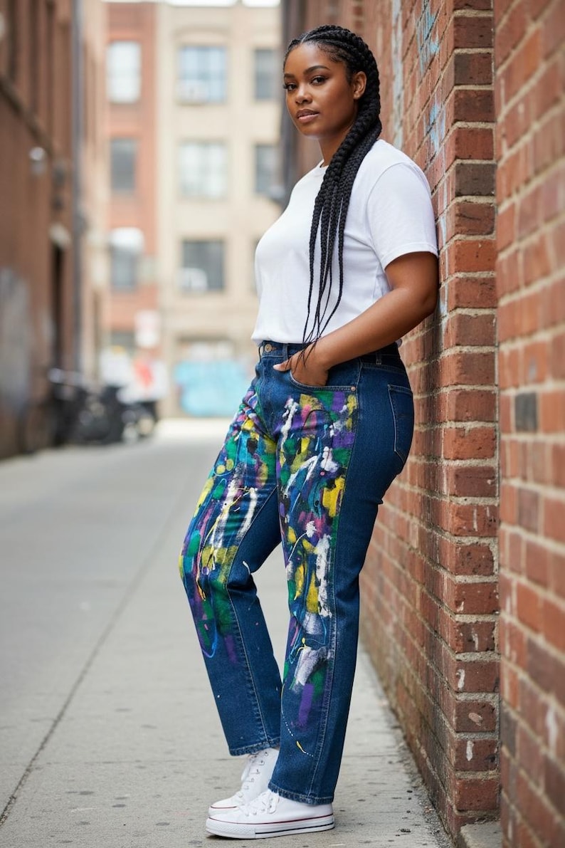 Pu&ograve; includere: Jeans in denim blu scuro con design a spruzzi di vernice colorata. I jeans sono abbinati a una maglietta bianca e a scarpe da ginnastica bianche. La modella &egrave; appoggiata a un muro di mattoni.