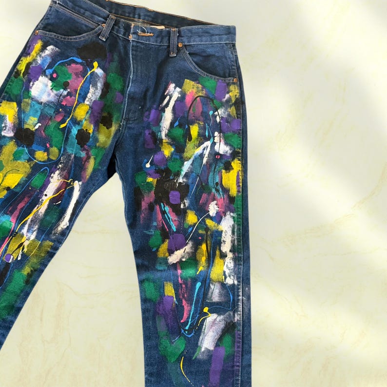 Pu&ograve; includere: Jeans di denim blu scuro con un colorato design astratto a spruzzi di vernice. La vernice comprende giallo, viola, bianco, verde e nero. I jeans hanno un classico design a cinque tasche.