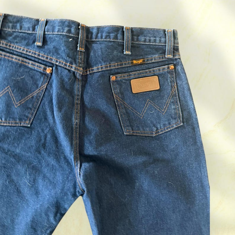 Pu&ograve; includere: Primo piano del retro di jeans in denim blu scuro. I jeans presentano due tasche posteriori con un caratteristico design a punto "W" e una toppa in pelle marrone con il logo "Wrangler". Le cuciture sono in un colore beige a contrasto.