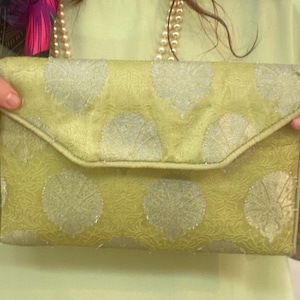 May include: A rectangular envelope-style clutch bag in a light lime green fabric featuring a repeating silver metallic leaf or fan-shaped pattern. The bag has a structured flap closure and a matching thin strap attached to the side.