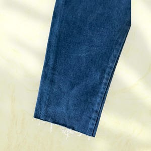 Pu&ograve; includere: Primo piano di una sezione di jeans blu. L'orlo &egrave; sfilacciato con fili bianchi. Il tessuto ha un aspetto strutturato ed &egrave; di una tonalit&agrave; media di blu.