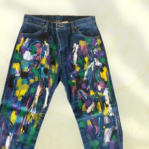 Pu&ograve; includere: Jeans di denim blu scuro con un design astratto e colorato a spruzzi di vernice. La vernice include giallo, verde, viola, rosa e bianco, creando un look unico e artistico. I jeans hanno un classico design a cinque tasche.