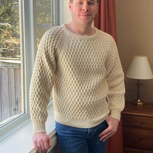 May include: A man wearing a cream knitted jumper with a textured pattern. He is standing in front of a window with a view of trees outside. He is wearing blue jeans and has a neutral expression on his face.