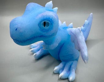 May include: A light blue dragon figurine with a detailed design. The dragon has a textured body, large eyes, and small wings. The figurine is made of a translucent material, giving it a soft appearance.