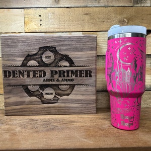 May include: A pink stainless steel tumbler with a silver design of a crescent moon, stars, and a castle. The tumbler is sitting on a wooden surface next to a wooden sign that says "Dented Primer Arms & Ammo Est. 2020".