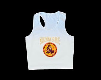 Può includere: Canotta bianca con orlo corto. La canotta presenta una grafica con la mascotte dell'Arizona State University, un diavolo dei cartoni animati, che tiene una palla da basket. Il testo "Arizona State" è stampato sopra la grafica.