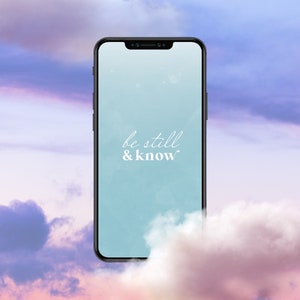 Op de afbeelding: Een smartphone met een blauwe achtergrond en de tekst "be still & know" op het scherm. De telefoon is omringd door wolken.