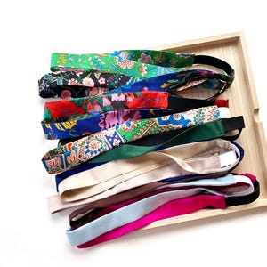 May include: A selection of patterned headbands in various colours, including green, blue, and pink, displayed on a wooden tray. The headbands feature floral and solid designs, with elasticated bands for a secure fit. The headbands are suitable for a range of hairstyles.