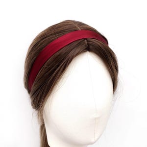 May include: A burgundy headband sits on a mannequin head with brown hair. The headband is a solid colour and seems to be made of a soft material. The hair is styled with a centre parting and falls down the back.