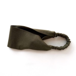 May include: An olive green headband with a twisted knot design. The headband features a wide, fabric band that tapers to a thin, elastic band at the back. The fabric appears to be a soft, natural material.