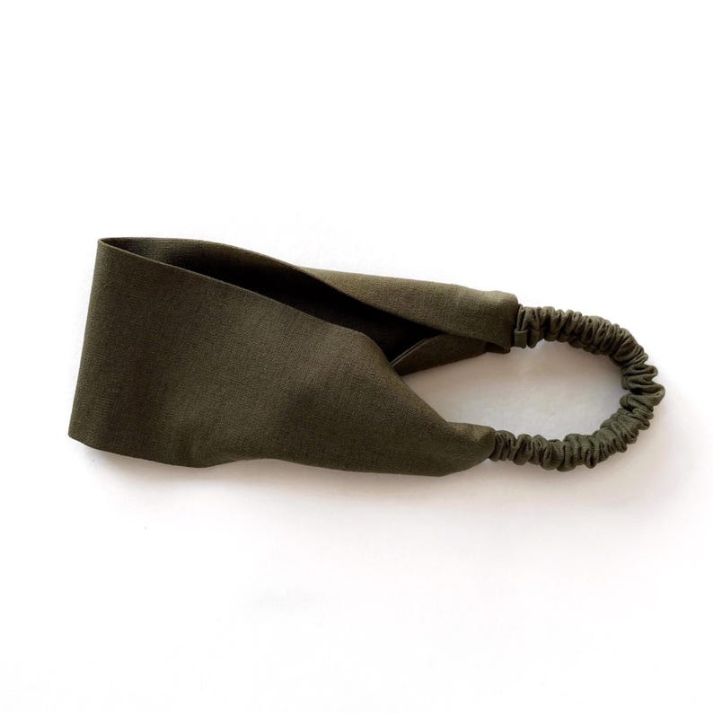 May include: An olive green headband with a twisted knot design. The headband features a wide, fabric band that tapers to a thin, elastic band at the back. The fabric appears to be a soft, natural material.