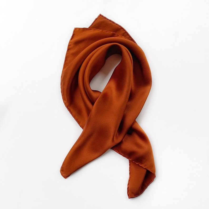 Pu&ograve; includere: Una sciarpa di seta color arancione bruciato, elegantemente drappeggiata su uno sfondo bianco. La texture liscia e il colore ricco della sciarpa sono evidenziati, mostrando il suo potenziale come accessorio di moda. La sciarpa quadrata &egrave; piegata e annodata.