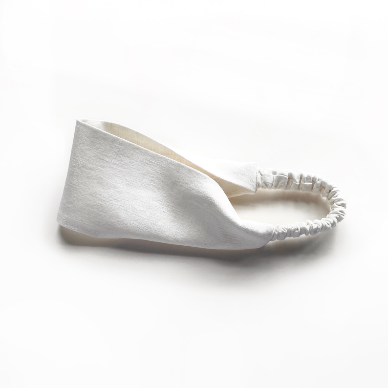 May include: A white headband with a textured fabric design. The headband features a wide, twisted front section and an elastic band at the back for a comfortable fit. This hair accessory is suitable for various hairstyles.