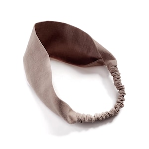 May include: A taupe-colored headband with a wide, knotted front and an elasticated back. The fabric has a textured appearance, and the headband is designed to be worn around the head. The neutral color makes it versatile for various outfits.