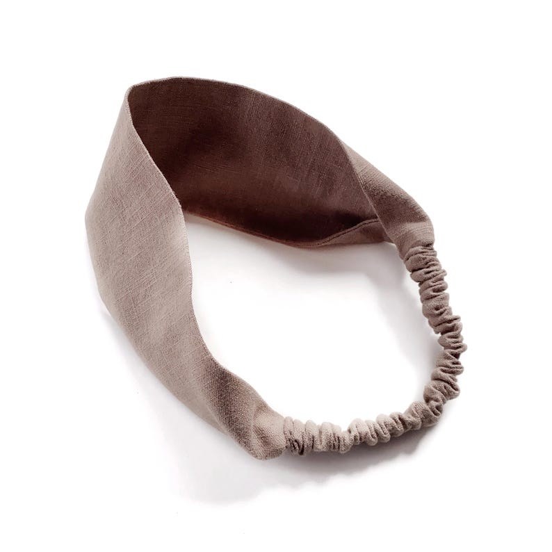 May include: A taupe-colored headband with a wide, knotted front and an elasticated back. The fabric has a textured appearance, and the headband is designed to be worn around the head. The neutral color makes it versatile for various outfits.