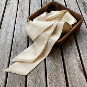 May include: A cream-colored satin ribbon is arranged in a wooden square bowl. The ribbon is folded and draped, with the ends extending out of the bowl. The wooden bowl has a dark brown finish and sits on a weathered wooden surface.
