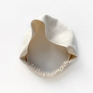 May include: A cream-colored linen headband with a wide, knotted design. The headband features an elastic band at the back for a comfortable fit. The fabric has a natural, textured appearance, and the overall style is simple and elegant.