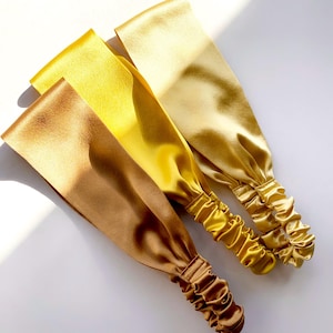Op de afbeelding: Drie gouden satijnen haarbanden, diagonaal geplaatst. De haarbanden hebben een gladde, glanzende textuur. De kleuren variëren van diep brons tot helder geel. De uiteinden van de haarbanden zijn met elastiek gerimpeld.