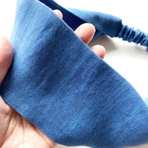 May include: A blue headband made of textured fabric. The headband has a wide, flat band and an elasticated section. The fabric is a solid, medium blue color. The headband is held in a hand.