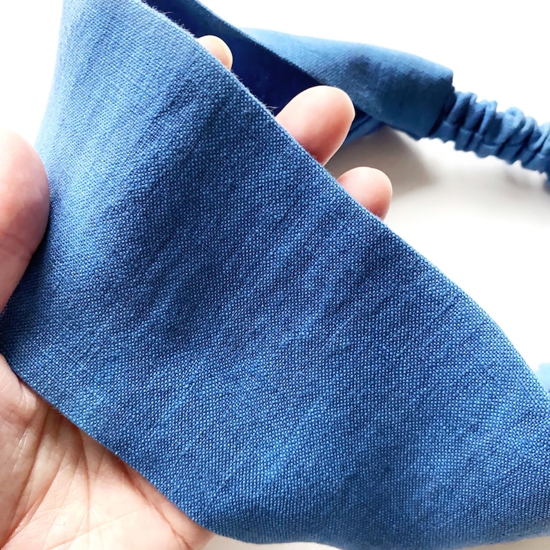 May include: A blue headband made of textured fabric. The headband has a wide, flat band and an elasticated section. The fabric is a solid, medium blue color. The headband is held in a hand.