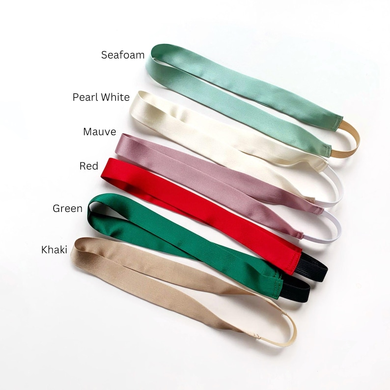 K&ouml;nnte beinhalten: F&uuml;nf Satin-Stirnb&auml;nder mit elastischen B&auml;ndern, in einem Stapel angeordnet. Die Farben sind als Seafoam, Pearl White, Mauve, Red, Green und Khaki beschriftet. Jedes Stirnband hat eine glatte, gl&auml;nzende Stoffoberfl&auml;che und ein flexibles, dehnbares R&uuml;ckenteil.