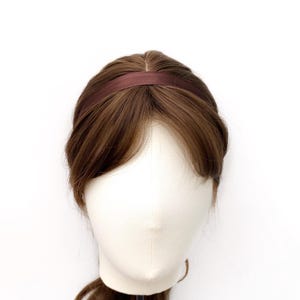 K&ouml;nnte beinhalten: Ein dunkelbraunes Satin-Haarband auf einem Schaufensterpuppenkopf mit hellbraunem Haar. Das Haarband hat eine glatte, einfarbige Stoffoberfl&auml;che und ein schmales, abgerundetes Profil, das oben auf dem Haar aufliegt und das Gesicht umrahmt.
