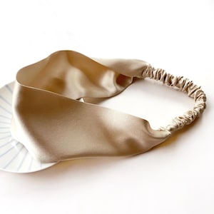 May include: A beige silk headband with an elasticated back. The headband is made of a soft, shiny material and is resting on a white plate with blue stripes.
