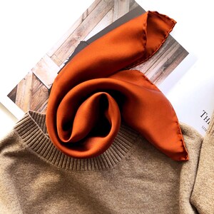 Pu&ograve; includere: Una sciarpa di seta arancione &egrave; drappeggiata su un maglione beige. La sciarpa &egrave; piegata e disposta per creare un effetto a spirale. Il maglione ha uno scollo a coste. Lo sfondo include una rivista e una superficie in legno.