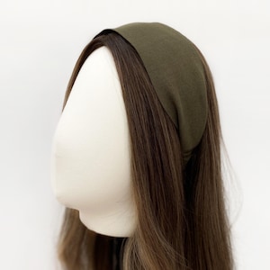 May include: An olive green headband is displayed on a mannequin head with long, brown hair. The headband is made of a soft fabric and is wide, covering the forehead. The hair is styled with a slight wave.