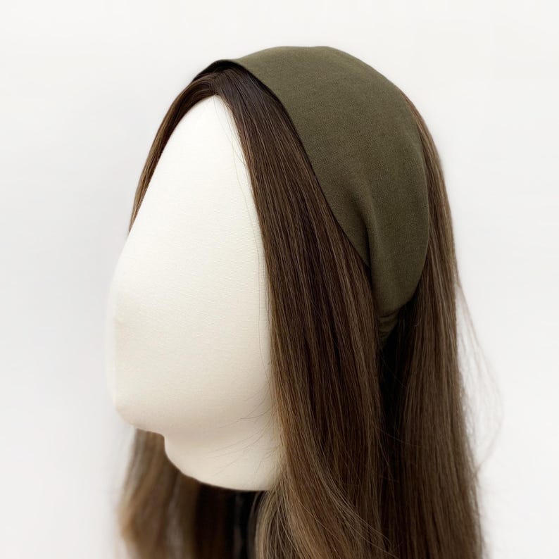 May include: An olive green headband is displayed on a mannequin head with long, brown hair. The headband is made of a soft fabric and is wide, covering the forehead. The hair is styled with a slight wave.