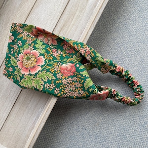 Op de afbeelding: Een brede stoffen haarband met een levendige groene achtergrond en een dicht bloemenpatroon van grote roze pioenen, kleinere lichtroze bloesems en groen blad. Het accessoire is voorzien van een elastisch gedeelte voor een goede pasvorm.