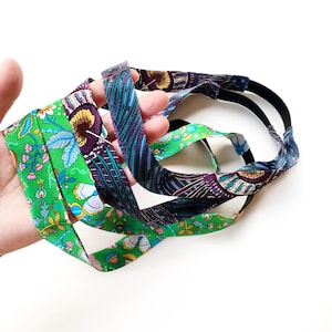 K&ouml;nnte beinhalten: Ein Set aus zwei Stoffstirnb&auml;ndern mit einem lebendigen Blumenmuster in leuchtendem Gr&uuml;n, Violett, Blau und Gelb. Jedes Stirnband besteht aus einem gemusterten Textilstreifen, der an einem einfarbigen schwarzen Gummiband befestigt ist.