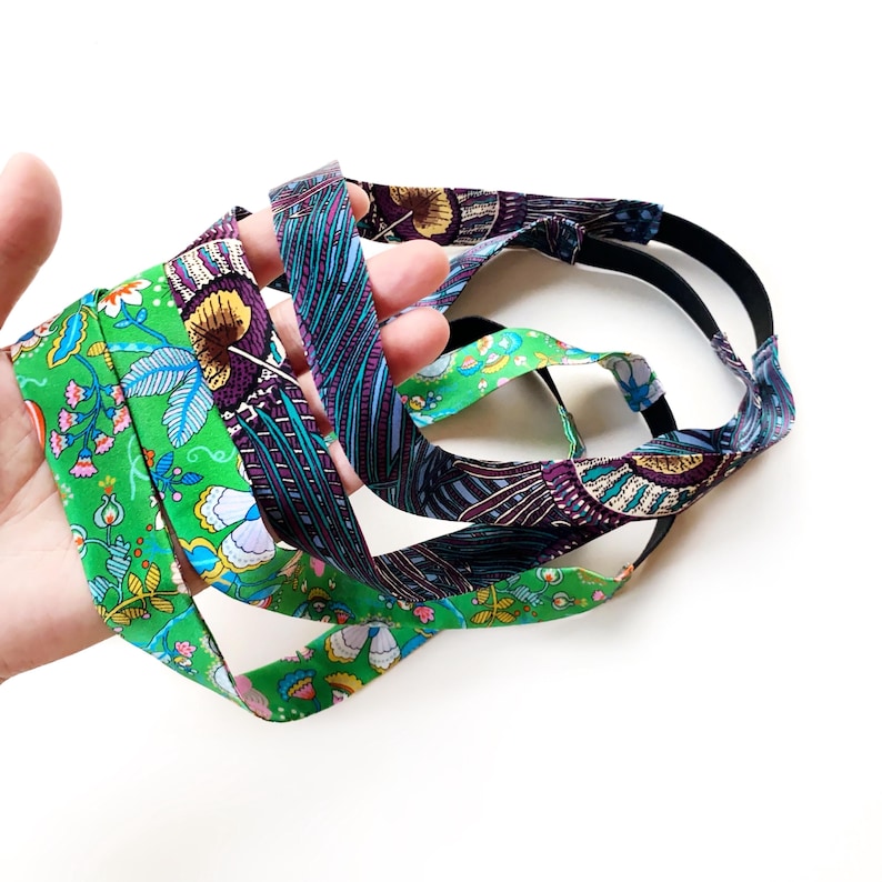 K&ouml;nnte beinhalten: Ein Set aus zwei Stoffstirnb&auml;ndern mit einem lebendigen Blumenmuster in leuchtendem Gr&uuml;n, Violett, Blau und Gelb. Jedes Stirnband besteht aus einem gemusterten Textilstreifen, der an einem einfarbigen schwarzen Gummiband befestigt ist.