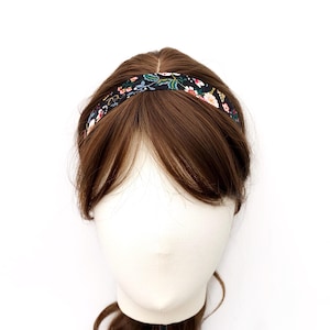 K&ouml;nnte beinhalten: Ein schmales Haarband mit schwarzem Hintergrund und einem bunten Blumenmuster aus wei&szlig;en, rosafarbenen und gr&uuml;nen botanischen Elementen. Das Haarband liegt auf dem Kopf einer braunhaarigen Schaufensterpuppe und ruht auf dem Haar.