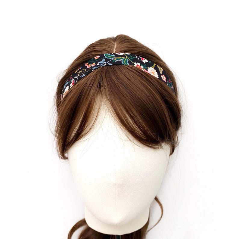 K&ouml;nnte beinhalten: Ein schmales Haarband mit schwarzem Hintergrund und einem bunten Blumenmuster aus wei&szlig;en, rosafarbenen und gr&uuml;nen botanischen Elementen. Das Haarband liegt auf dem Kopf einer braunhaarigen Schaufensterpuppe und ruht auf dem Haar.