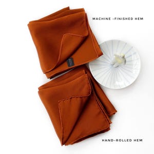 Pu&ograve; includere: Due tovaglioli piegati color ruggine con orli rifiniti a macchina e arrotolati a mano sono esposti. Accanto ai tovaglioli c'&egrave; un piattino bianco con strisce blu e una statuina decorativa. Il testo "MACHINE - FINISHED HEM" e "HAND-ROLLED HEM" &egrave; visibile.