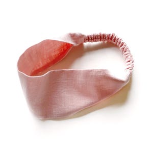 May include: A light pink headband with a twisted knot design. The headband features a wide, textured band and an elasticated back for a comfortable fit. The interior of the band is a slightly darker shade of pink.