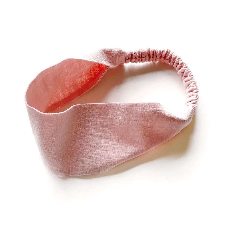 May include: A light pink headband with a twisted knot design. The headband features a wide, textured band and an elasticated back for a comfortable fit. The interior of the band is a slightly darker shade of pink.