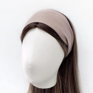 May include: A taupe-colored headband made of soft fabric. The wide headband is shown on a mannequin head with long brown hair. The headband is a solid color and appears to be made of linen or a similar material.