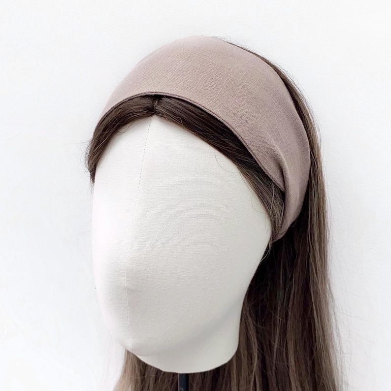 May include: A taupe-colored headband made of soft fabric. The wide headband is shown on a mannequin head with long brown hair. The headband is a solid color and appears to be made of linen or a similar material.