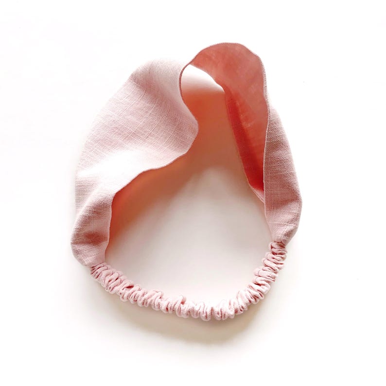 May include: A light pink linen headband with an elastic band. The headband is twisted at the top and has a soft, textured appearance. The elastic band is a matching pink color.