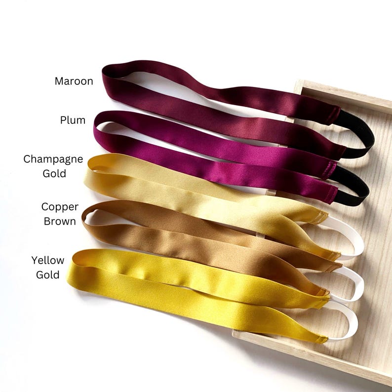 K&ouml;nnte beinhalten: F&uuml;nf Satin-Stirnb&auml;nder mit elastischen B&auml;ndern, beschriftet als Maroon, Plum, Champagne Gold, Copper Brown und Yellow Gold. Jedes Stirnband hat eine glatte, gl&auml;nzende Oberfl&auml;che und ein flexibles Schlaufendesign f&uuml;r einen sicheren Halt.
