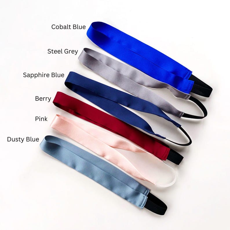 K&ouml;nnte beinhalten: Eine Sammlung von sechs Satin-Stirnb&auml;ndern, die diagonal angeordnet und mit Cobalt Blue, Steel Grey, Sapphire Blue, Berry, Pink und Dusty Blue beschriftet sind. Jedes Stirnband besteht aus einem glatten, gl&auml;nzenden Stoffband mit schwarzem Gummizug.