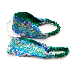 May include: Two headbands with a floral and bird pattern in shades of blue, green, pink, and yellow. The interior of the headband is green, and the elastic band is a matching green color. The headbands are on a white background.