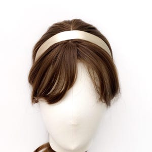 K&ouml;nnte beinhalten: Ein glattes, einfarbiges champagnerfarbenes Stirnband auf einem Schaufensterpuppenkopf mit braunem Haar. Das Stirnband hat ein schlichtes, geschwungenes Design mit matter Oberfl&auml;che, das sicher auf dem Kopf sitzt und das braune Haar sowie den Pony umrahmt.