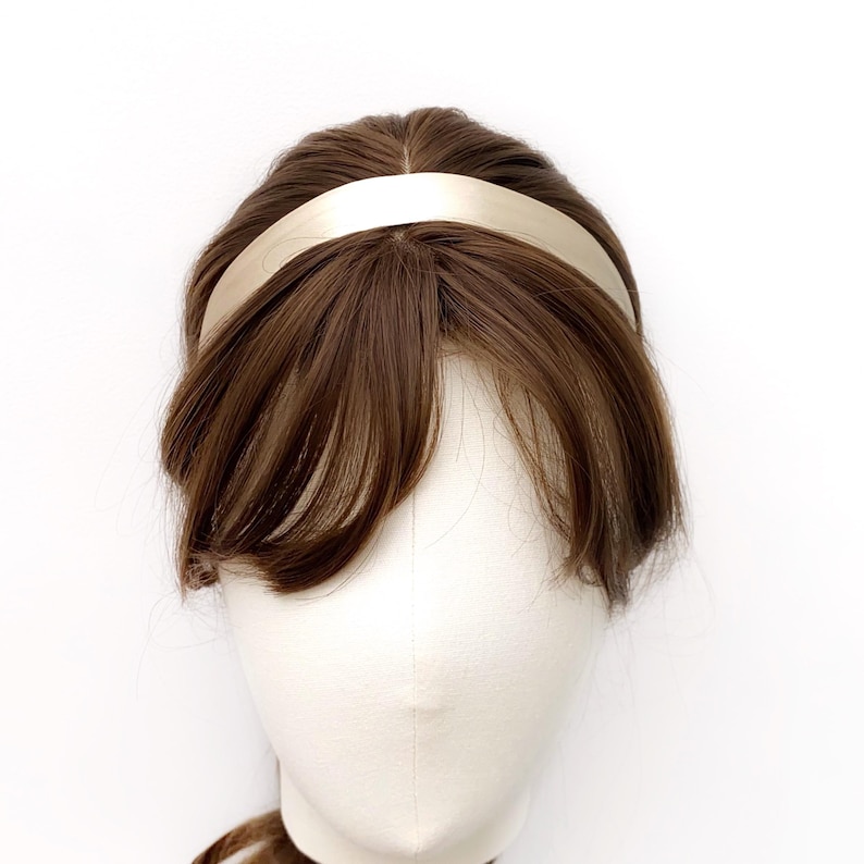 K&ouml;nnte beinhalten: Ein glattes, einfarbiges champagnerfarbenes Stirnband auf einem Schaufensterpuppenkopf mit braunem Haar. Das Stirnband hat ein schlichtes, geschwungenes Design mit matter Oberfl&auml;che, das sicher auf dem Kopf sitzt und das braune Haar sowie den Pony umrahmt.
