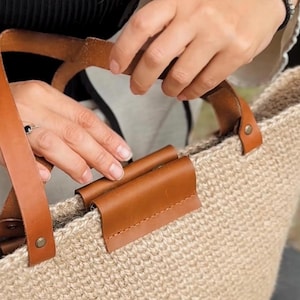 Op de afbeelding: Een beige geweven draagtas met bruine leren handvatten en accenten. De tas heeft een stevig, open ontwerp, ideaal voor het dragen van diverse items. De leren handvatten zijn bevestigd met metalen klinknagels, wat een rustieke charme toevoegt.