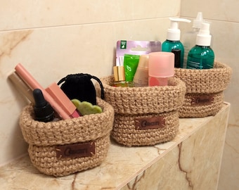 May include: Three woven jute baskets of varying sizes, each filled with beauty products. The baskets have a natural, light brown colour and are labelled with a small leather tag that reads "Clean". The baskets are on a marble shelf.