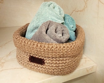 May include: A brown crocheted basket with a leather label sits on a white shelf. The basket is filled with rolled up towels, one light blue and two grey.