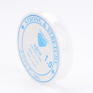 Peut inclure: Une bobine de fil élastique transparent étiquetée "STRONG & STRETCHY" avec un logo en forme de diamant et le texte "水の縦 CRYSTA LINE 1.0" et "ポリウレタン 100%"
